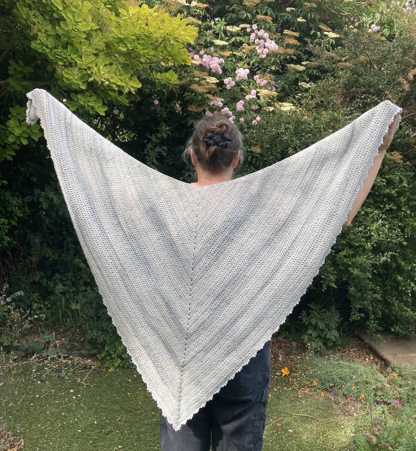 A pale blue and cream crochet triangular shawl held open outdoors against a flowering hedge