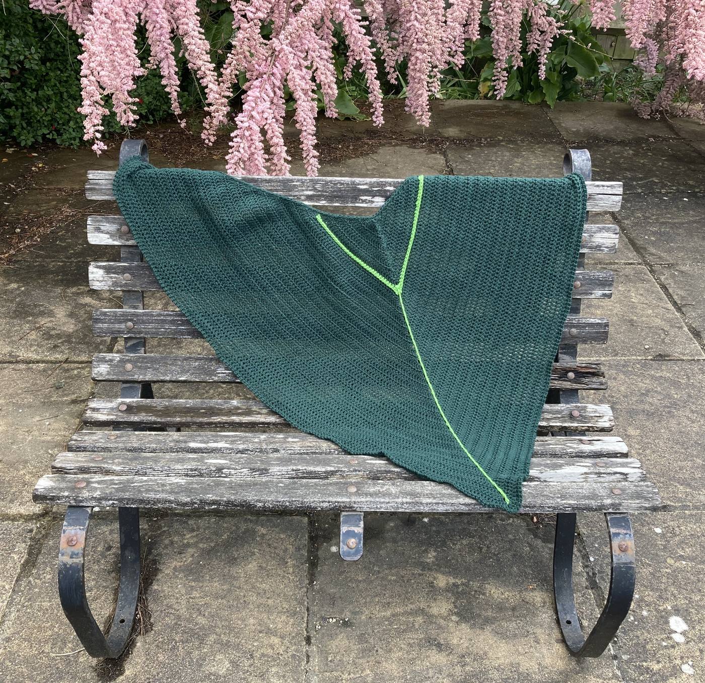 A deep green crochet shawl shaped like a leaf, draped over a weathered garden bench, with a bright lime green seam line
